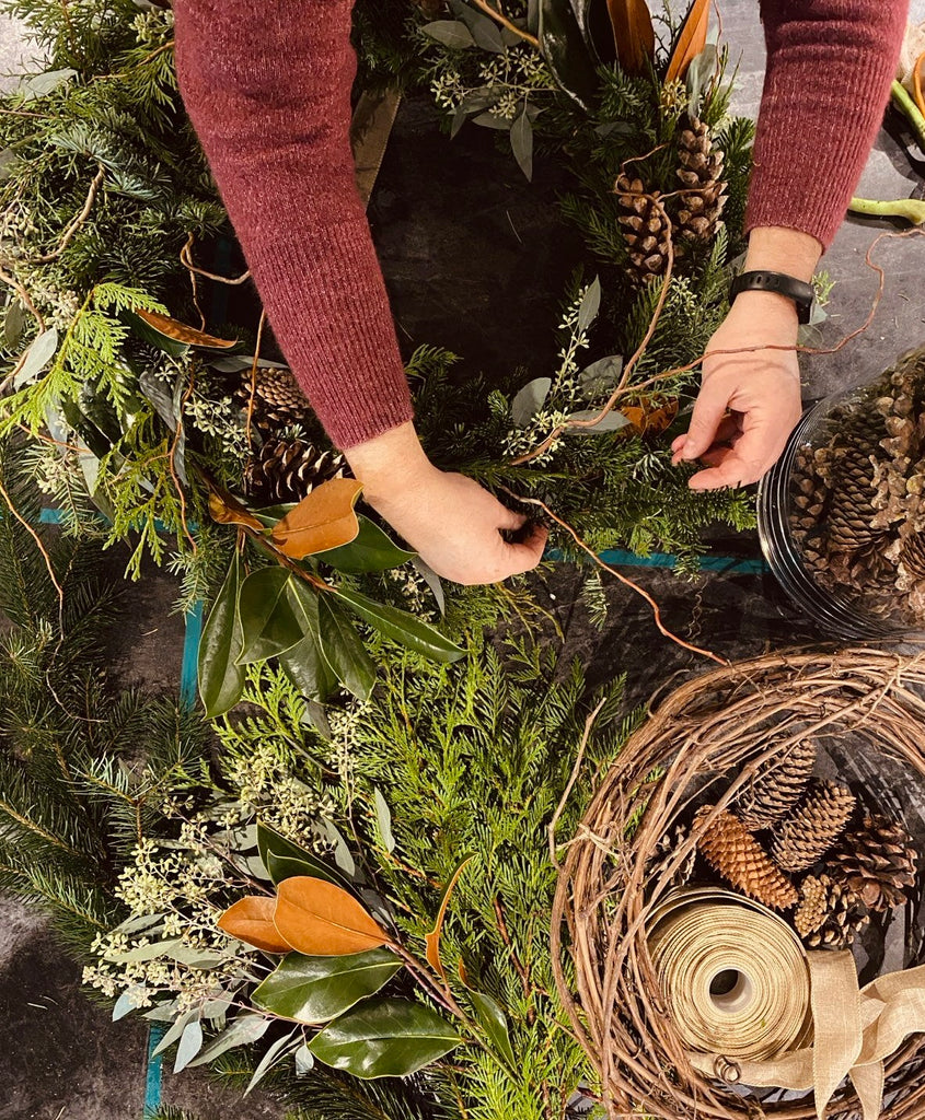 Mains d'une participante en train de préparer une couronne de Noël avec des matériaux naturels, lors de l'atelier du 30 novembre et 7 décembre 2024. Vue prise de haut, mettant en valeur la créativité.