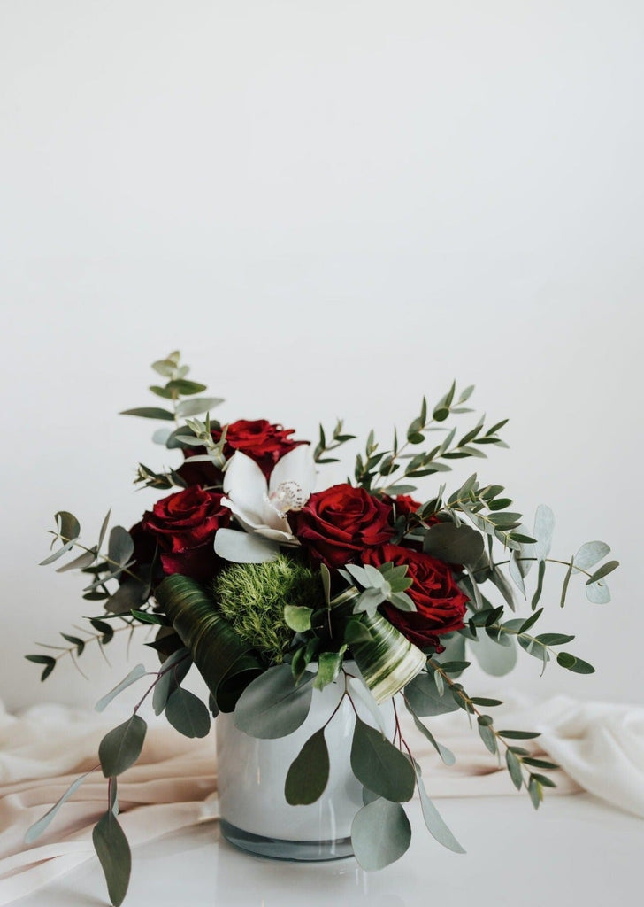 Composition florale "À la Folie" avec roses rouges, orchidée blanche et feuillage naturel dans un vase blanc.