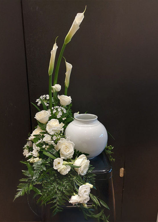 Arrangement floral funéraire blanc avec roses, callas et verdure entourant une urne blanche.