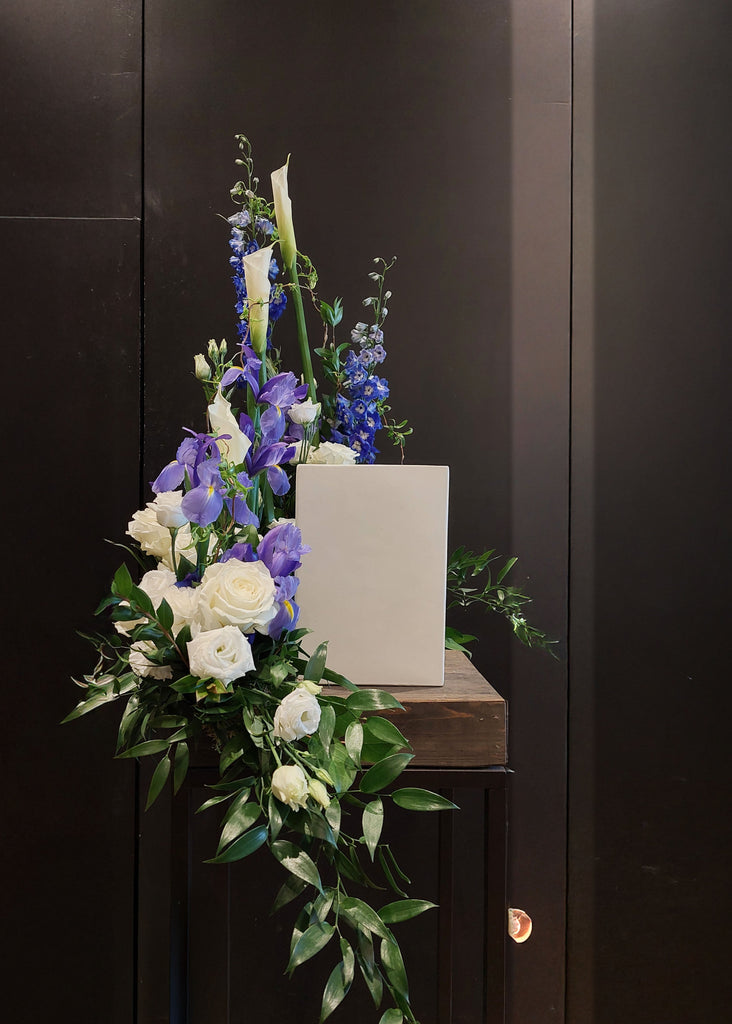 Arrangement floral funéraire en bleu et blanc avec roses, callas et delphiniums entourant une urne blanche.