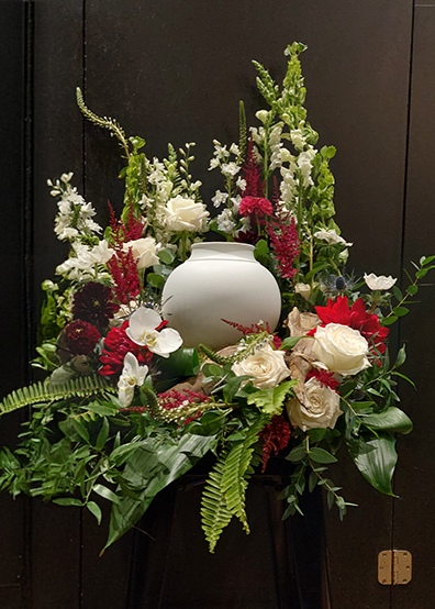 Arrangement floral funéraire rouge et blanc avec roses, orchidées et feuillage entourant une urne blanche.